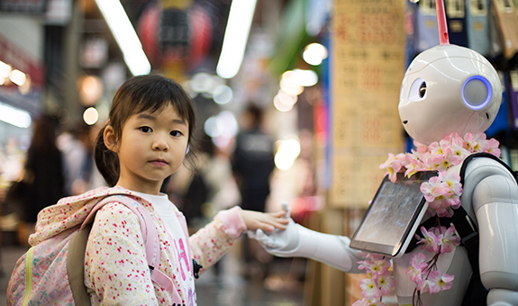 Ein in rosa gekleidetes asiatisches Mädchen, das in die Hand von einem menschenähnlichen weißen Roboter hält und in die Kamera guckt.
