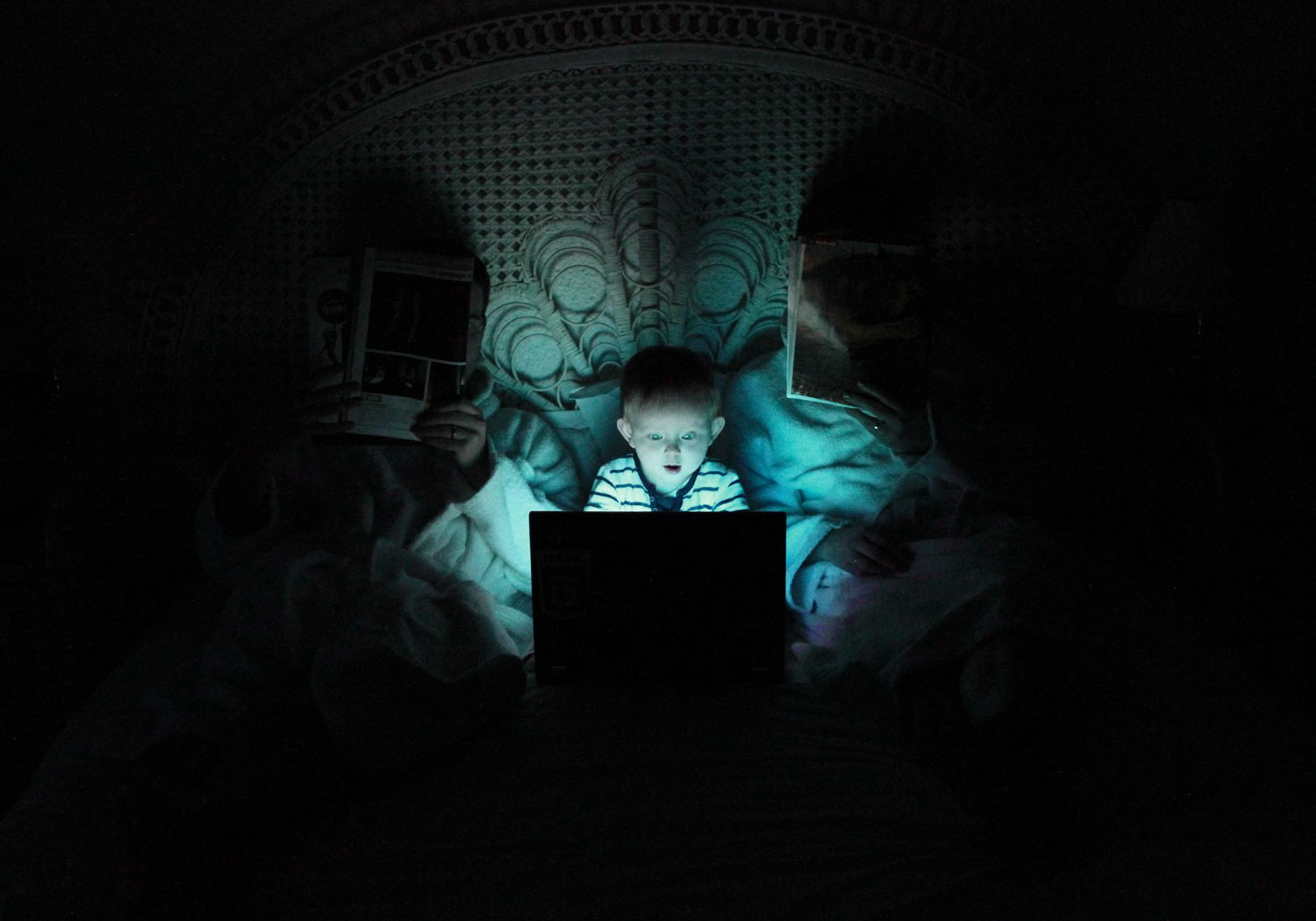 Baby sitzt im dunkeln zwischen Eltern im Bett und starrt fasziniert auf einen blau leuchteten Computer Bildschirm.