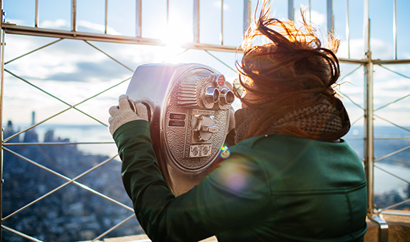 Frau mit braunen Haaren, dunkel grüner Jacke und Schal ist auf dem Empire State Building in New York und schaut durch ein Fernglas.