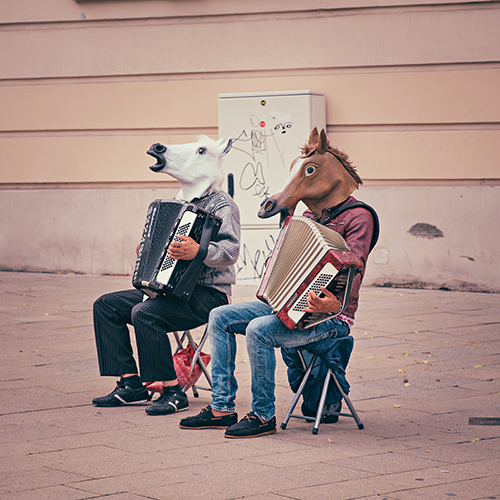 Zwei Musiker mit Akkordions und Pferdekopfkostümen sitzen auf Klappstühlen in der Straße und spielen Musik.