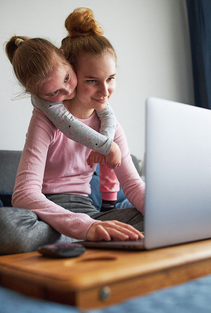 Ein jugendliches Mädchen sitzt am Computer während ihre kleine Schwester ihr über die Schulter guckt.