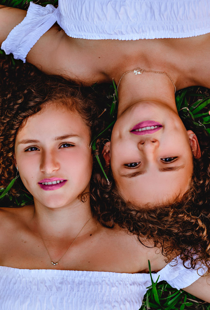 Zwillinger mit lockigen Haar und pinken Lippenstift liege nebeneinander auf einer Wiese und gucken in die Kamera.