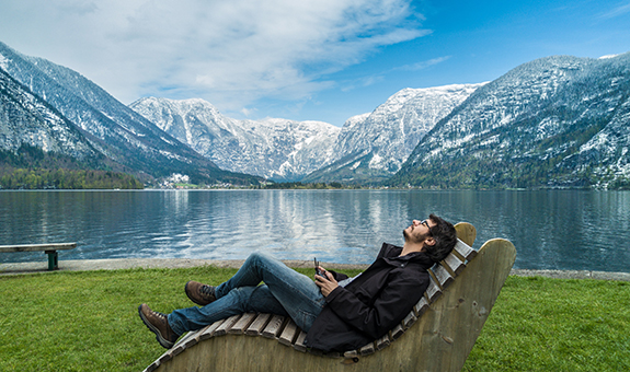 Dunkelhaariger Mann in Outdoor Klamotten liegt au f einer Holzbank vor einem See und einer Berglandschaft im Hintergrund und schaut in den Himmel.